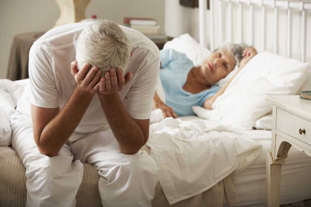 Worried,Senior,Man,Sits,On,Bed,Whilst,Wife,Sleeps