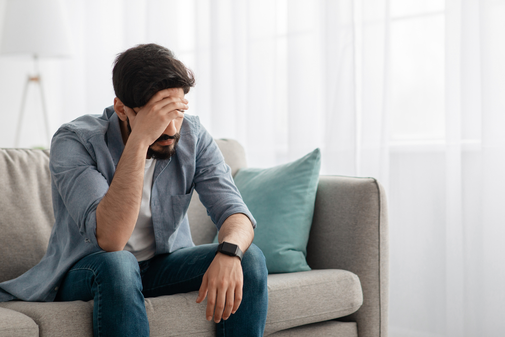 young man sitting on couch with head resting on his hand upset